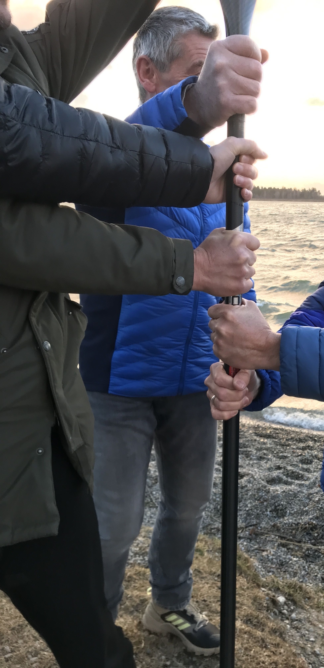 Männerhände an einem Paddelstiel. Die Männer stehen auf dem Strand eines Sees.