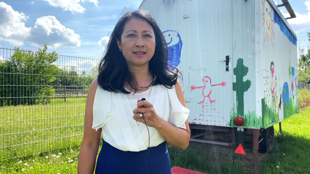 Mai Dang-Goy steht mit Mikrofon vor einem von Kindern bemalten Bauwagen. Sie gibt ein Interview.